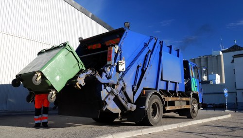 A large commercial rubbish collection truck, painted blue with black lower sections, is positioned on a paved outdoor area adjacent to a white industrial building. The truck's rear hydraulic compactor mechanism is raised, in the process of emptying a green wheeled waste bin into the container, with the lid open, showing a partially filled interior. The waste bin is made of plastic, has a textured surface, and is mounted on small wheels. A worker dressed in red trousers and a high visibility safety vest is standing nearby, guiding or monitoring the disposal process. The scene is illuminated by daylight, with a clear blue sky and some scattered clouds in the background, indicating an outdoor waste collection or rubbish removal activity typical of services provided by companies like Commercial Waste Removal Hanwell in the local area near London. Visible structures in the background include factory-type buildings and utility poles, situating the scene within an industrial or commercial environment relevant to rubbish or waste disposal services.