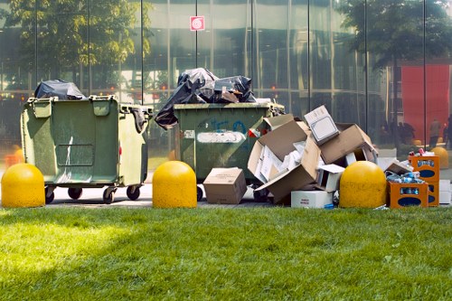 A scene showing three large wheelie bins positioned on a paved area adjacent to a grassy verge, likely outdoors in an urban or suburban environment. The left bin is green with a closed lid, and the right one appears to be green as well, both showing signs of use and some wear. In front of and around these bins, there are various discarded cardboard boxes, some flattened and others loosely assembled, along with black plastic bags that seem partially open or sagging. The cardboard boxes vary in size and are scattered, with some stacked or leaning against the bins. There is also a small orange plastic crate containing miscellaneous rubbish on the right side. The background features a reflective glass façade of a building with visible trees and a red structure, indicating proximity to a commercial or office area. The scene is illuminated by natural daylight, with shadows cast by the bins and rubbish, suggesting a clear or partly cloudy sky. This setup aligns with rubbish collection or waste removal services in Hanwell, providing a detailed visual reference for a commercial waste removal context.