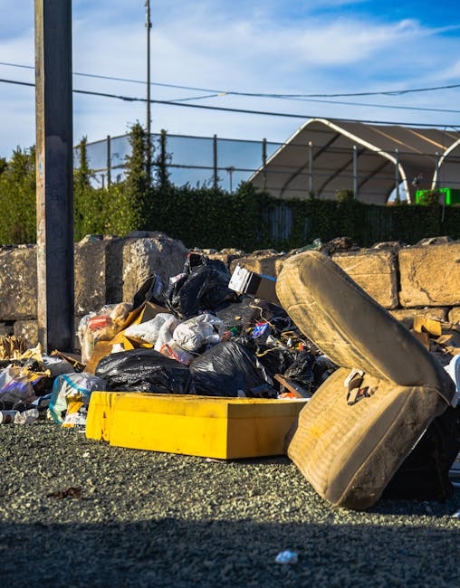 A pile of discarded rubbish is shown on a gravel surface, consisting of black plastic bin bags, cardboard and paper waste, and a large, worn, beige car seat with fabric upholstery lying on its side. T