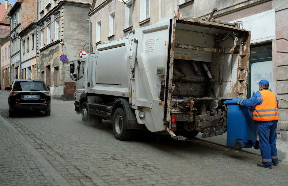 A narrow urban alleyway filled with a large pile of mixed waste and discarded materials, including cardboard boxes, black plastic bags, and other refuse, all stacked against a weathered wall topped wi